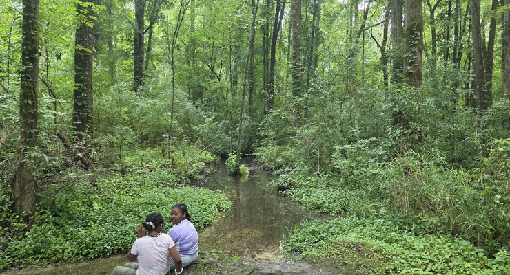 Two children sitting beside the wooded stream at God's Acre Healing Springs surrounded by lush green trees in Blackville SC