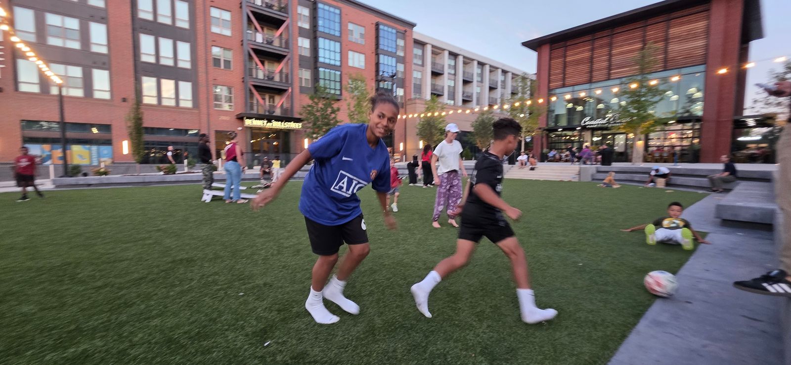 A boy and girl chase a soccer ball across the turf lawn at Gather COLA